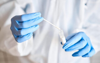 A lab worker in a white coat and blue rubber gloves holds a sample swab over a clear tube